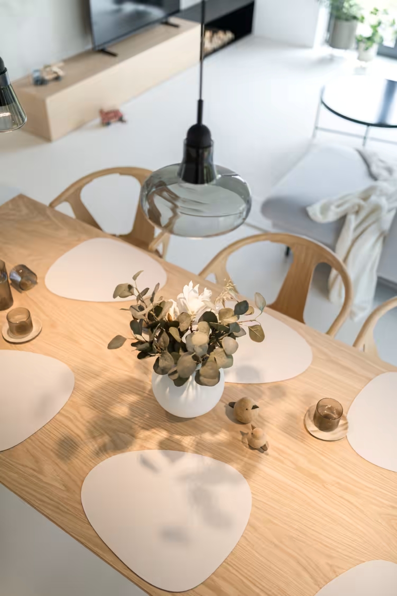 A wooden dining table with six uniquely shaped white place mats, a white vase with green foliage and white flowers at the centre, and candles, in a modern, minimalistic living space.