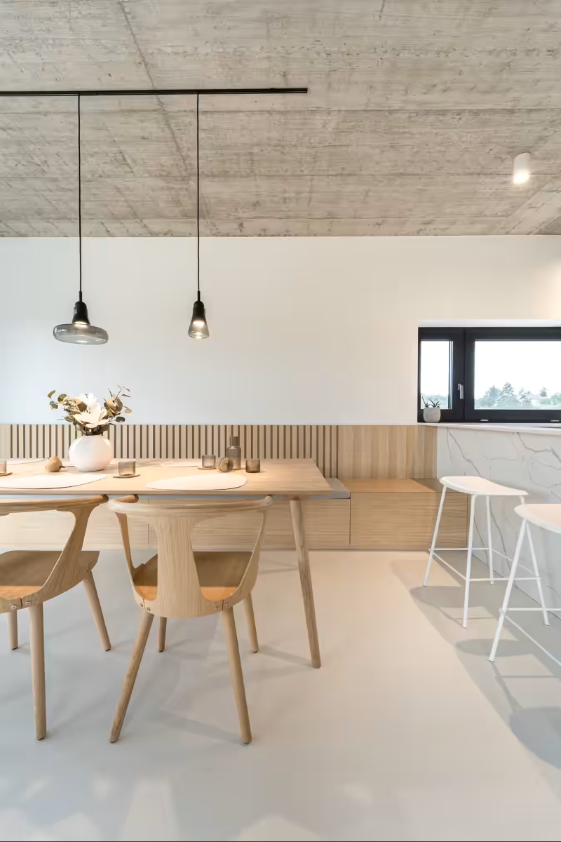 Minimalist dining area with light wooden table and chairs, modern pendant lights, a vase with flowers, a small window, and a white bar stool near a marble worktop against a neutral, contemporary background.