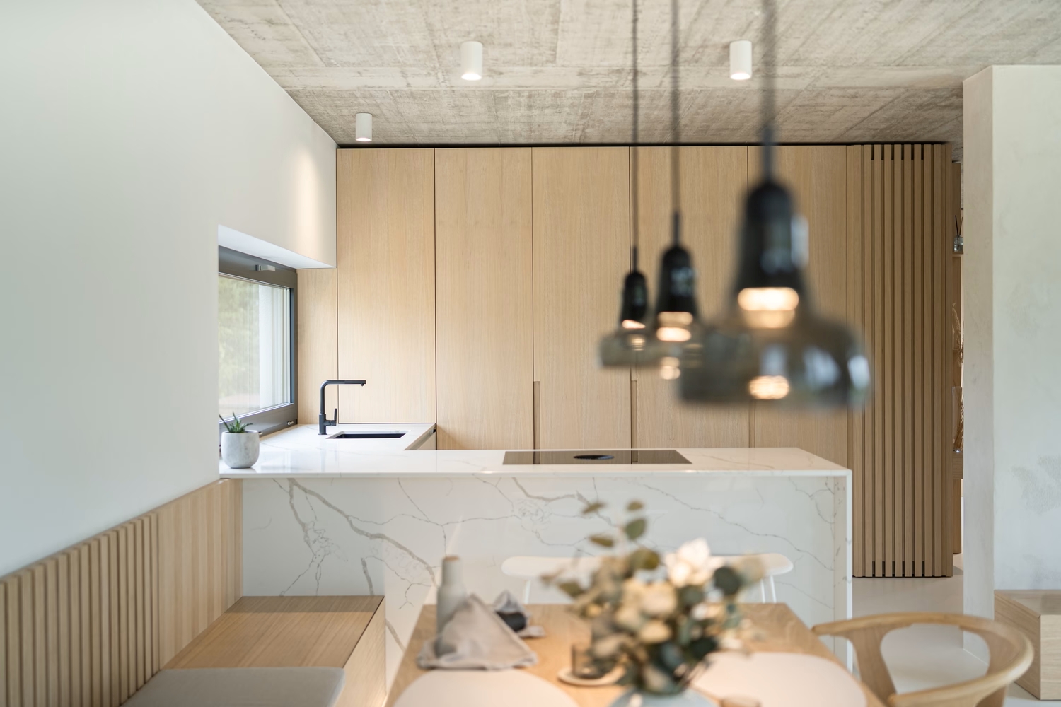 Minimalist kitchen with light wood cupboards, a white marble island, black pendant lights, and a small dining table with a flower centrepiece. A window above the sink lets in natural light.