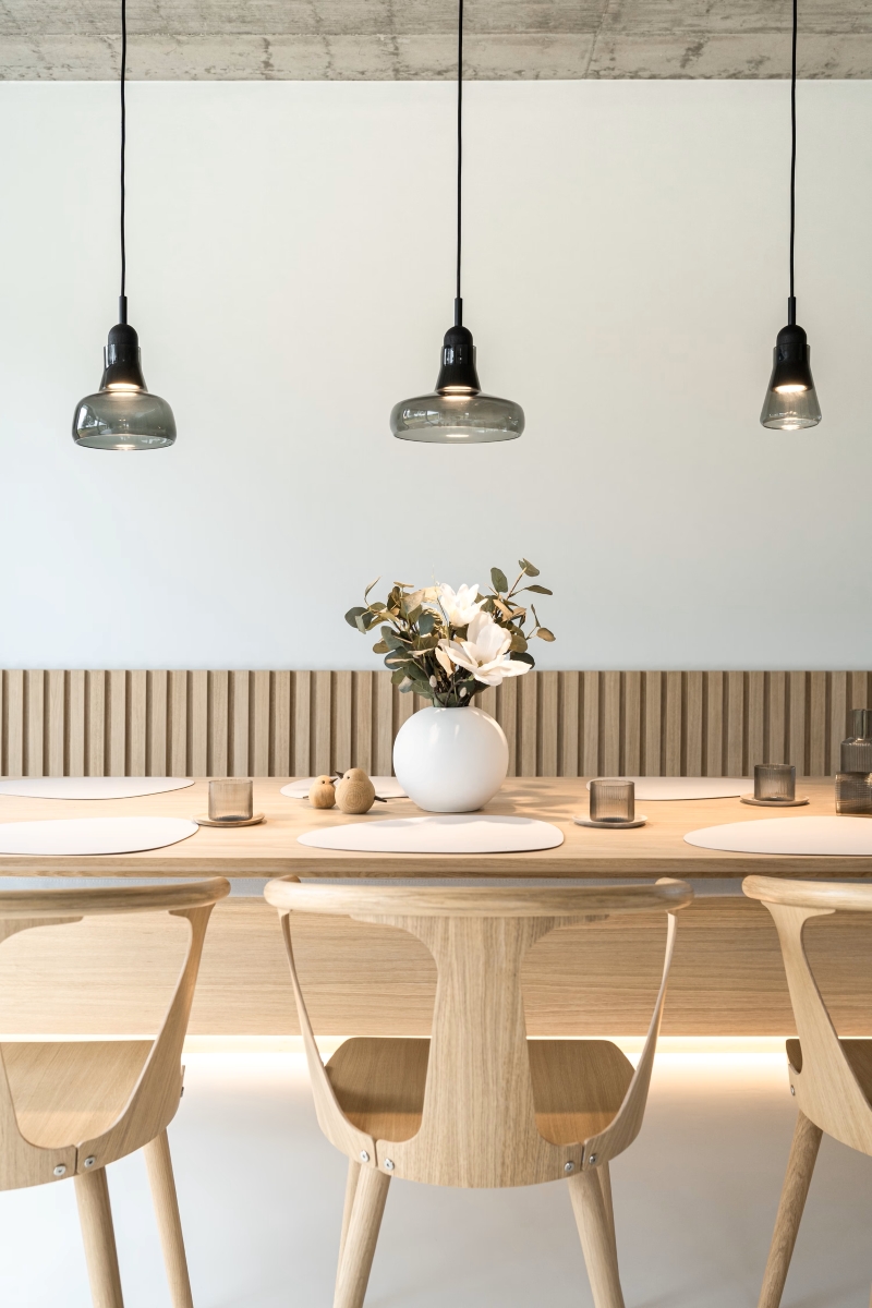 Minimalist dining room with three wooden chairs, a light wooden table, white placemats, a white vase with flowers, and three hanging pendant lights against a simple, modern background.