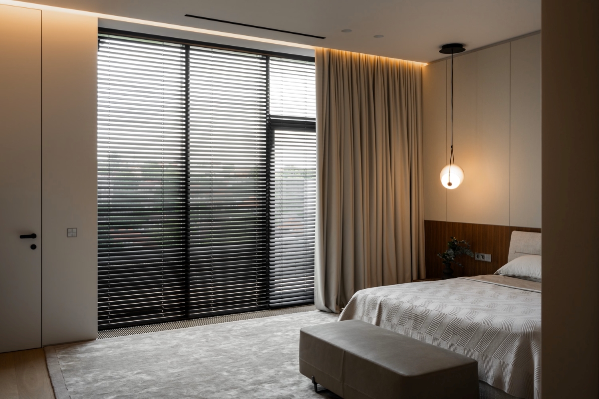 A modern bedroom with a large window covered by horizontal blinds and beige curtains, a bed with light bedding, a bench at the foot, and a hanging round light fitting in the corner.