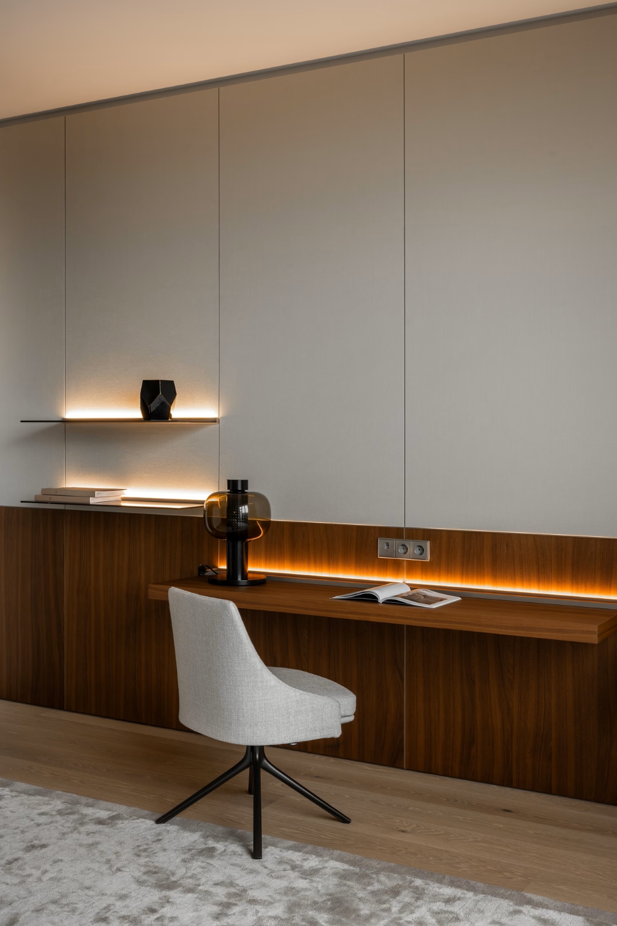 Minimalist home office with a built-in wooden desk, modern grey chair, book, lamp, and decorative objects. Soft LED lighting illuminates the shelves and wall panels, creating a warm, elegant atmosphere.