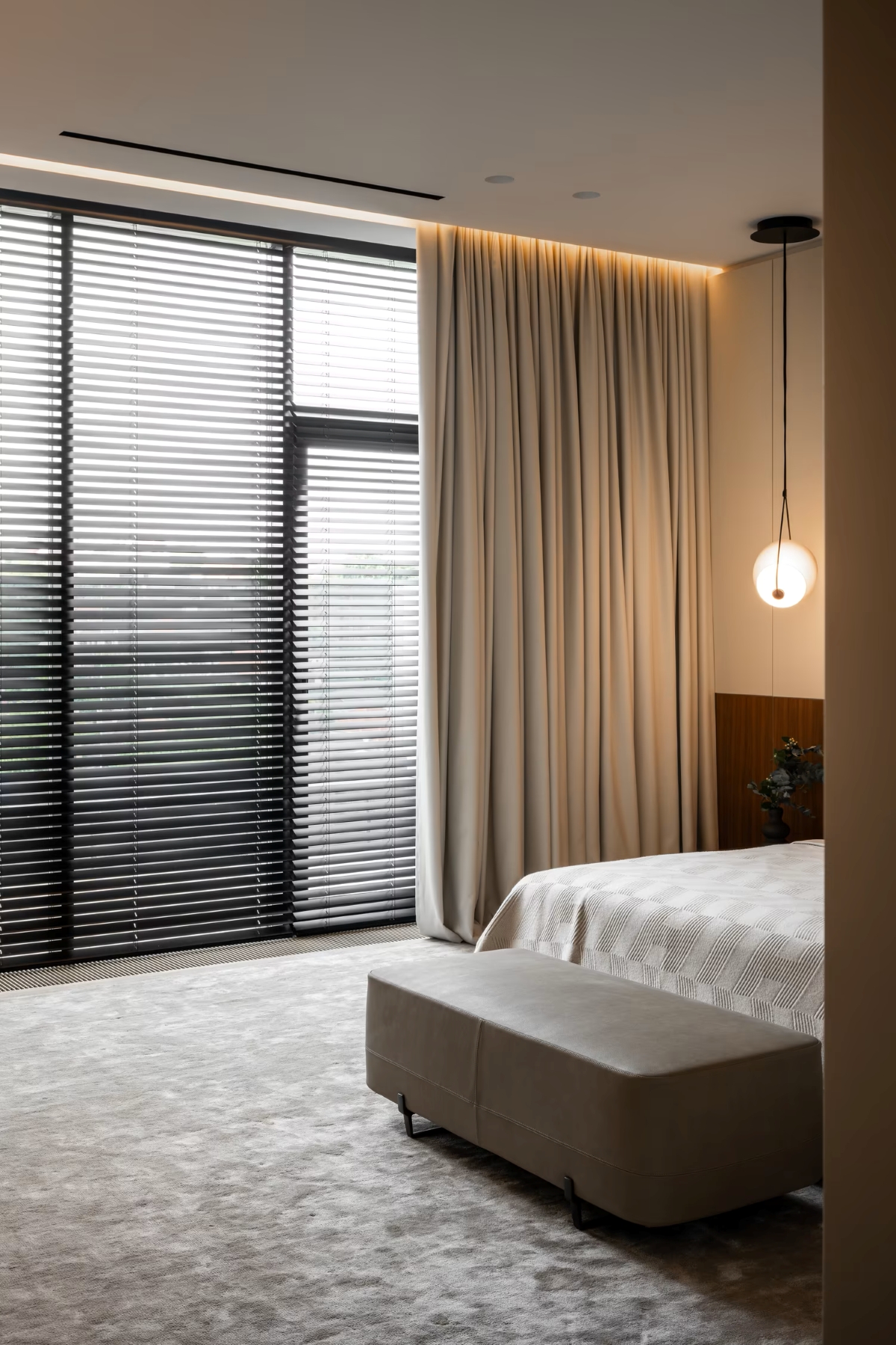 A modern bedroom with floor-to-ceiling windows covered by black blinds and beige curtains, a bed with neutral bedlinen, a cushioned bench at the foot, and a round pendant light hanging beside the bed.