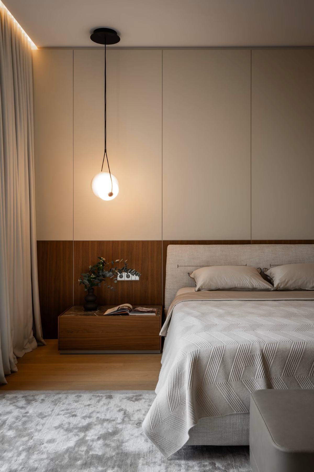 A modern, minimal bedroom with a beige and wooden feature wall, a pendant light, a neatly made bed with light bedding, a wooden bedside table with books and decor, and sheer curtains beside a soft grey rug.