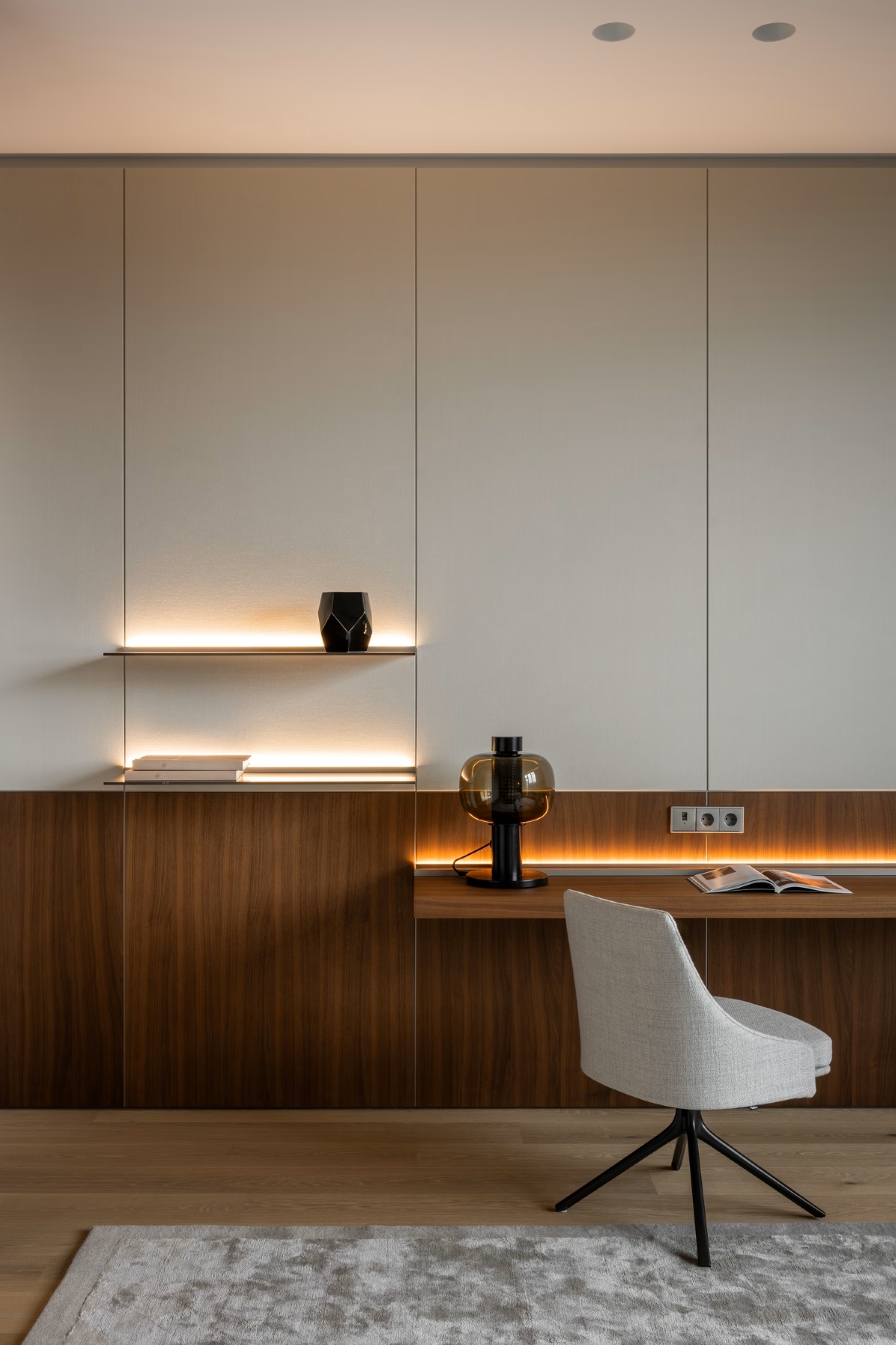 Minimalist home office with a white swivel chair, wooden desk, open book, table lamp, decorative vase, and wall-mounted shelves with soft lighting. Neutral tones and clean lines create a calm atmosphere.