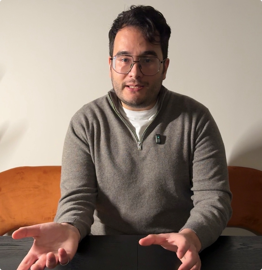 A man with dark hair and glasses, wearing a grey jumper, is sitting at a table with his hands open and facing upwards. He appears to be speaking or explaining something, and is seated against a plain wall.