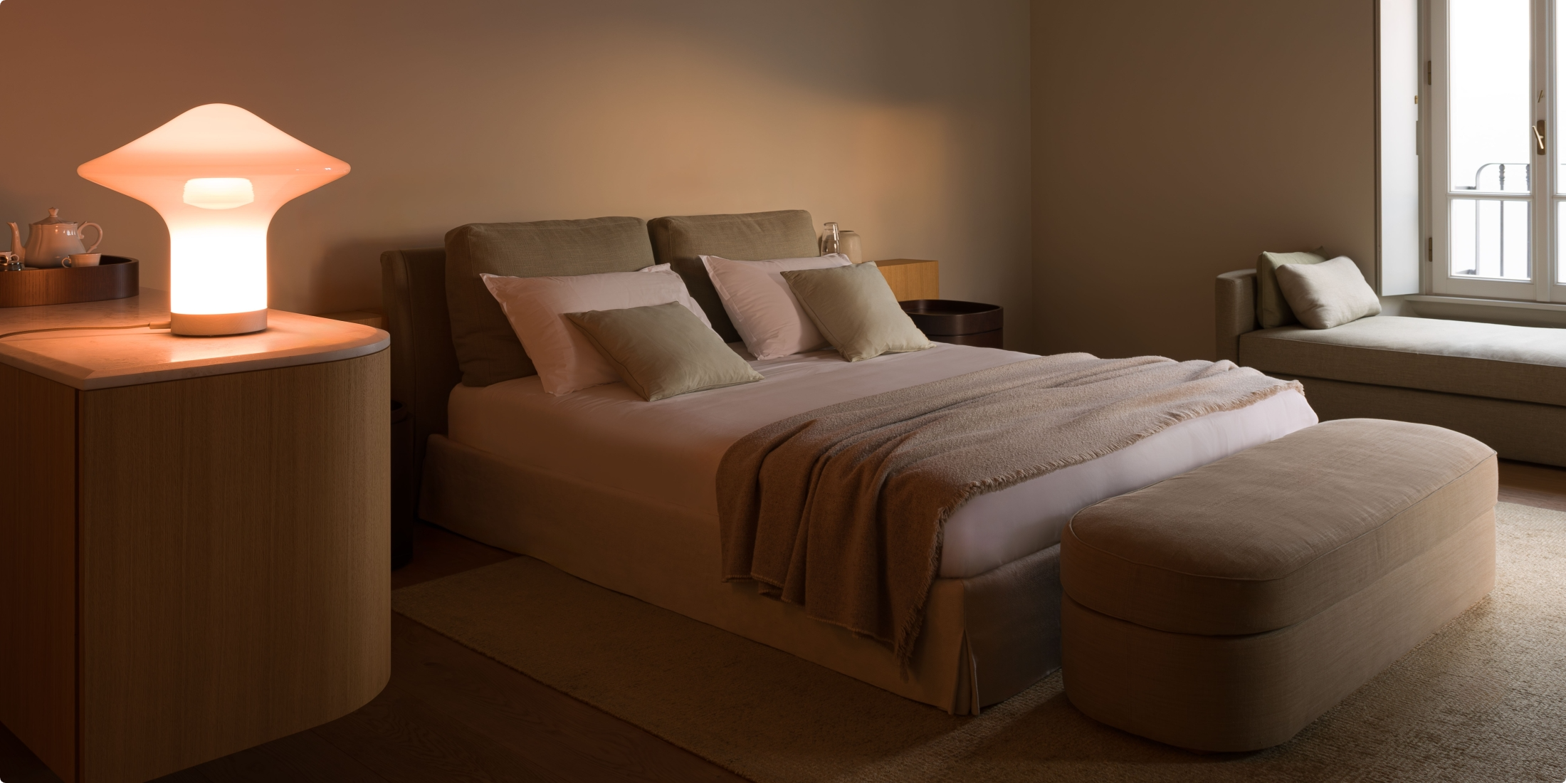 A cosy, softly lit bedroom with a double bed, neutral bedding, two pillows, a beige ottoman at the foot, a sideboard with a glowing lamp, and a window seat by a large window. The décor is minimalist and modern.