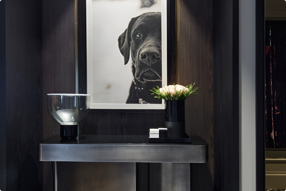 A modern console table with a clear lamp, a vase of white flowers, and books, beneath a large framed black-and-white photo of a black dog, set against a dark wall.