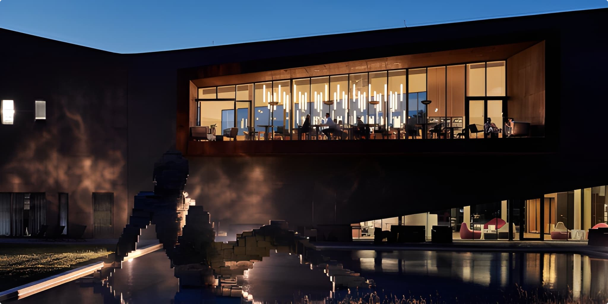 A modern building with a large, glass-walled upper storey glows warmly at dusk. Below, a geometric sculpture stands in a reflective pool surrounded by soft outdoor lighting.