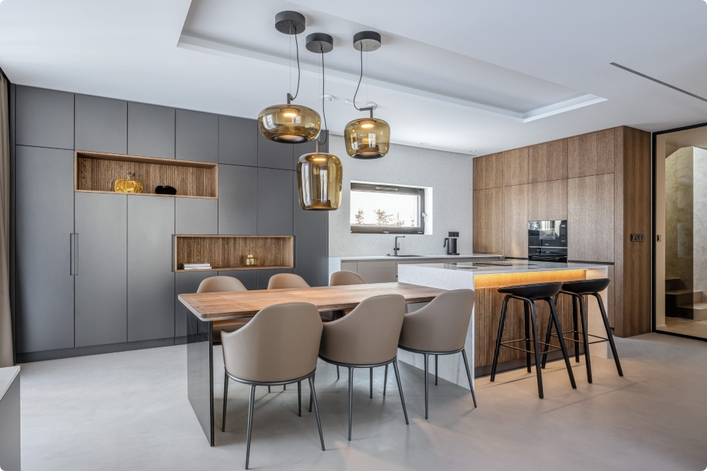 Modern kitchen and dining area with wood and grey cabinets, a wooden dining table, beige chairs, black bar stools at a white island, and three glass pendant lights. A small window lets in natural light.