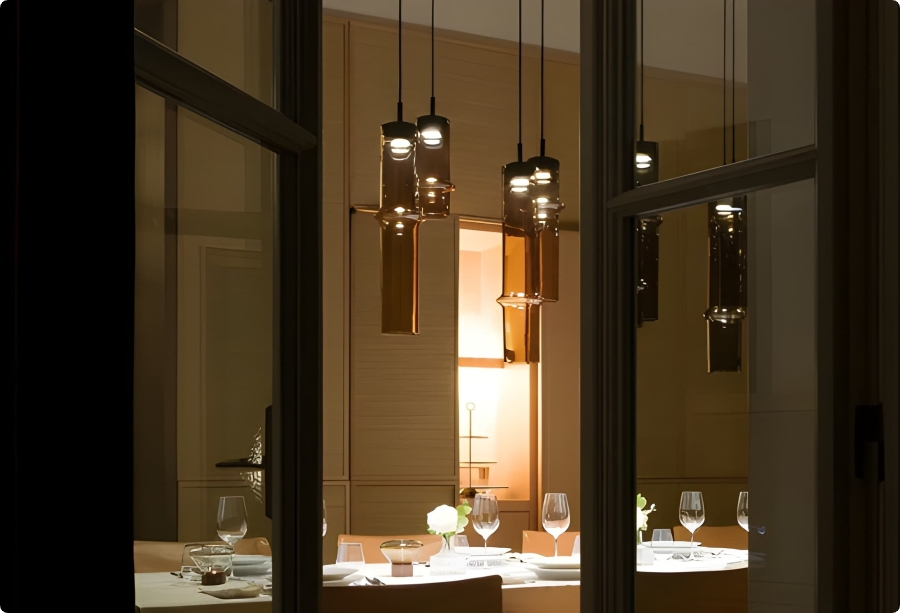 A modern dining room viewed through a window, featuring a set table with wine glasses, plates, and white flowers. Several stylish pendant lights hang above the table, illuminating the elegant, softly lit interior.