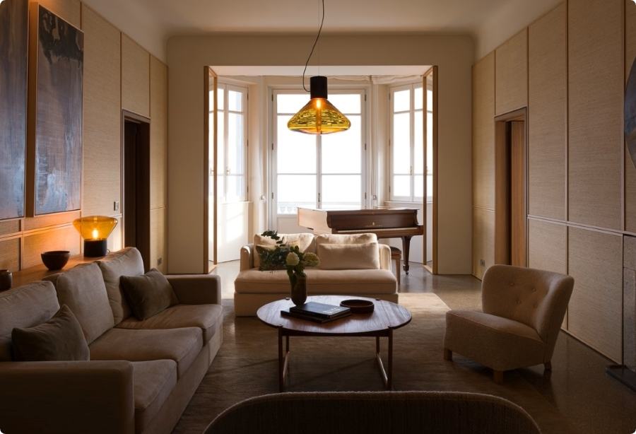 A cosy living room with beige sofas, a round wooden coffee table, flowers, and soft lighting. Large windows in the background let in natural light, and a grand piano sits near the sunny window nook.