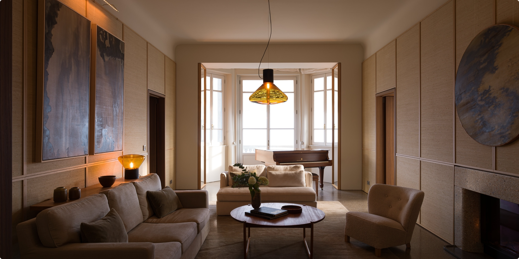 A cosy, modern living room with beige sofas, a round coffee table, large abstract artwork, a piano by tall windows, and warm lighting from a pendant lamp and table lamp.