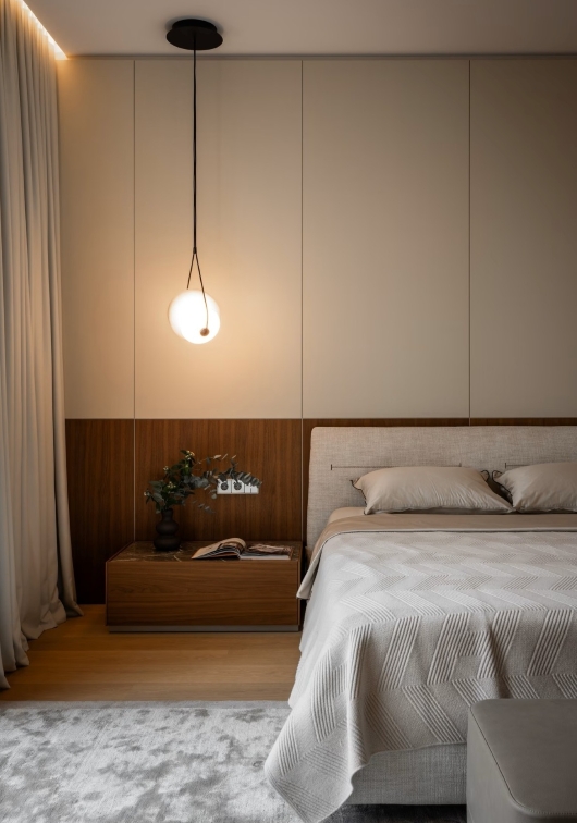 A modern bedroom with a beige and wood feature wall, a bed with a textured blanket, and a bedside table holding a vase and books. Overhead, a Brokis round pendant light glows as soft natural light enters through sheer curtains.