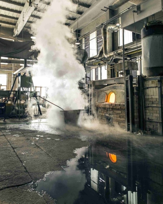 A person near a large industrial furnace stirs a metal container, creating a thick cloud of steam. The scene is inside a factory with concrete floors, puddles, and pipes visible overhead.