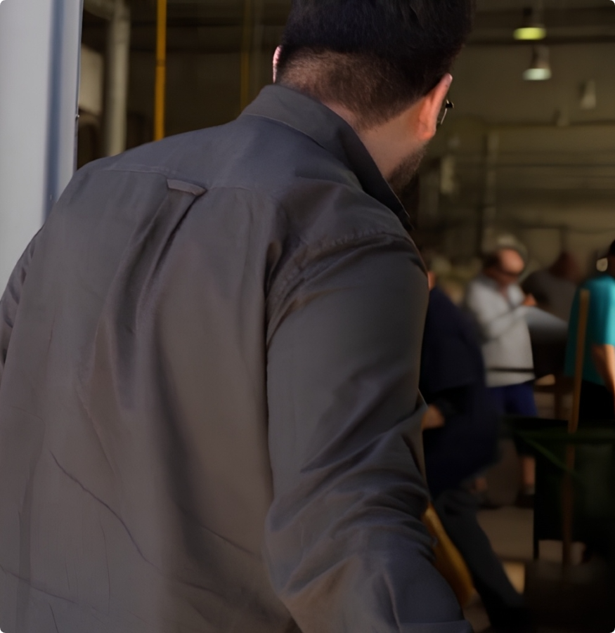 A man with short dark hair, wearing glasses and a grey jacket, is seen from behind as he enters a busy indoor workspace illuminated by stylish Brokis lighting, with several people in the background.