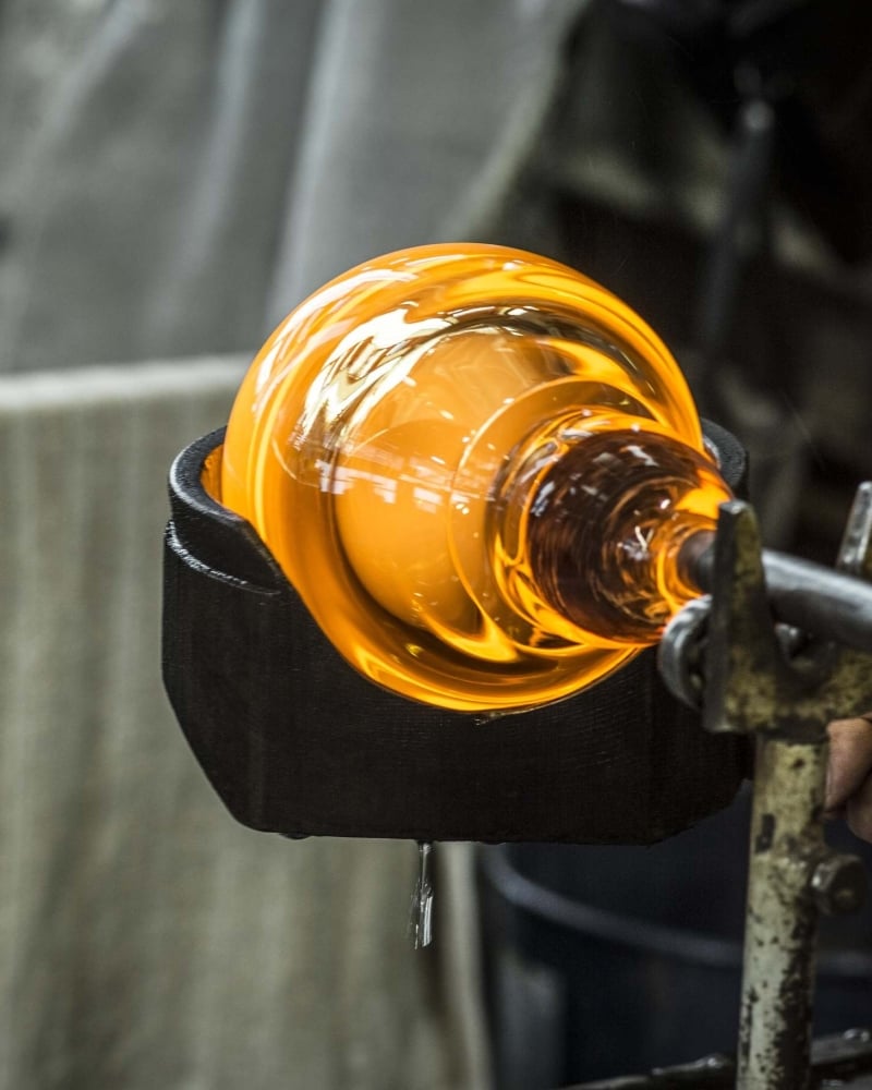 A glowing orange-hot glass object is being shaped with a tool during the glassblowing process, with the molten glass attached to a metal pipe—craftsmanship often seen in Brokis lighting creations.