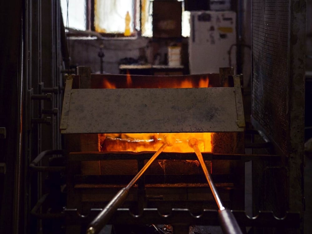Two metal rods are inserted into a glowing, fiery Brokis furnace in a dimly lit industrial workshop, with flames visible inside the furnace and various equipment in the background.