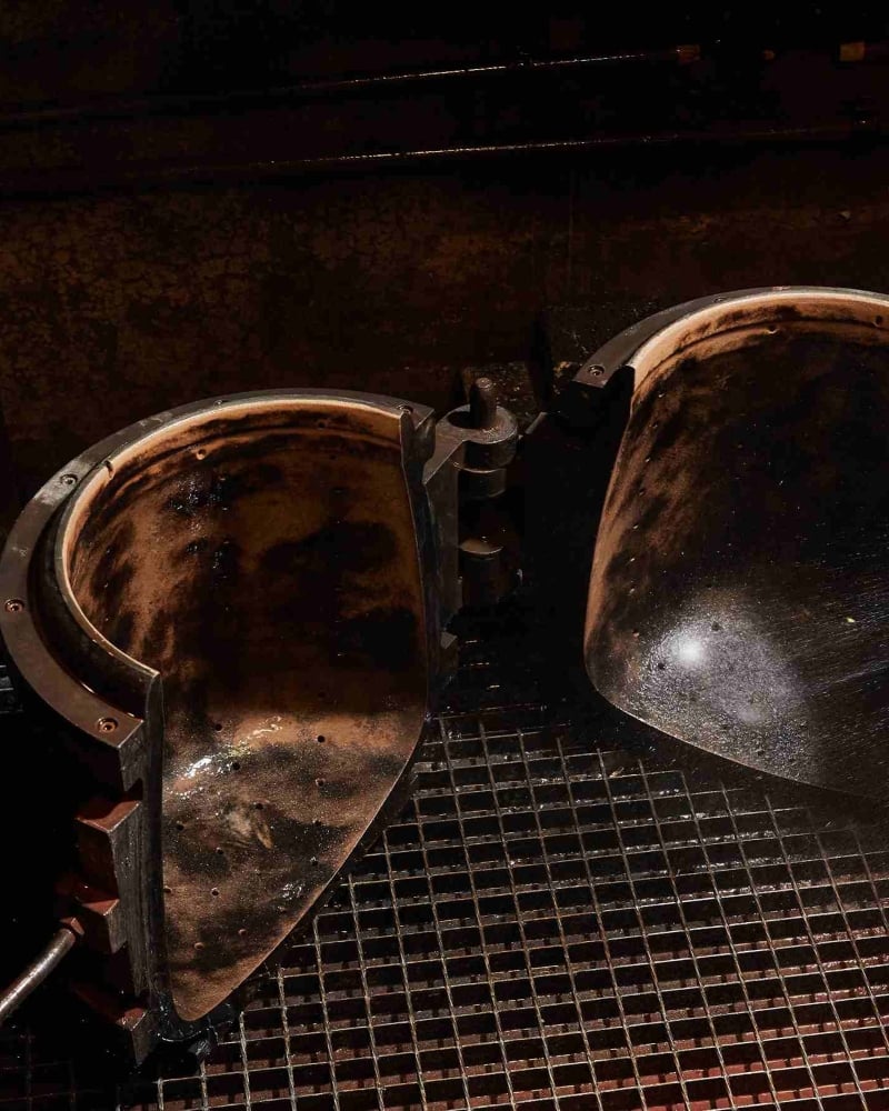 A large, open, rounded metal mould with a worn, darkened interior sits on a metal grate, illuminated under warm lighting in an industrial setting, reminiscent of Brokis’ signature craftsmanship.