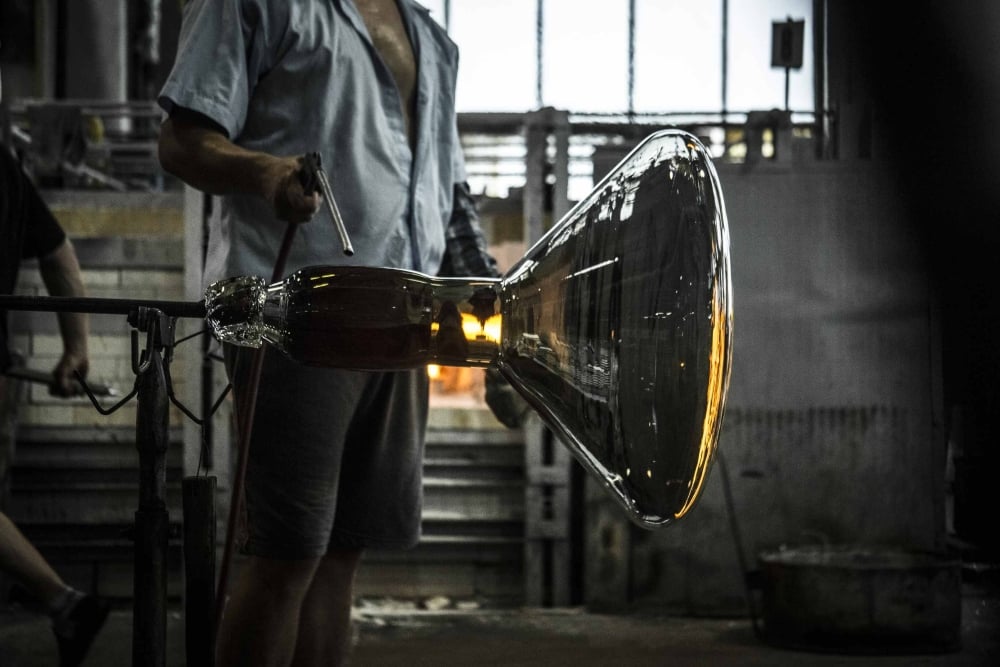 A person shapes a large piece of molten glass using a metal tool in an industrial workshop, with the glowing glass reflecting light—a process reminiscent of Brokis’s renowned glass artistry.