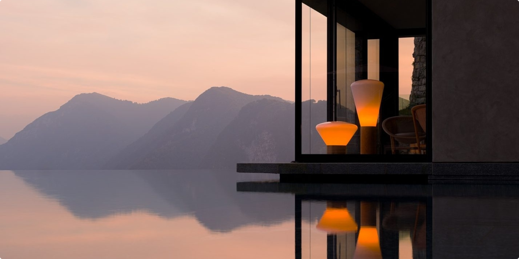 A modern house with large glass windows glows warmly at dusk, featuring elegant Brokis lighting and overlooking calm water that reflects the light. Mountains are visible in the background under a soft pink and orange sky.