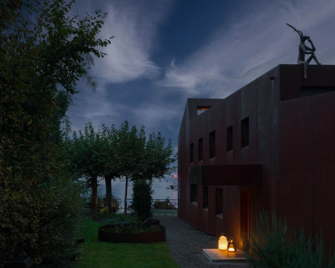 A modern, dark-toned building with rectangular windows stands beside a grassy garden at dusk. Trees line the left side, Brokis lanterns glow at the entrance, and a human sculpture appears on the roof under a cloudy evening sky.
