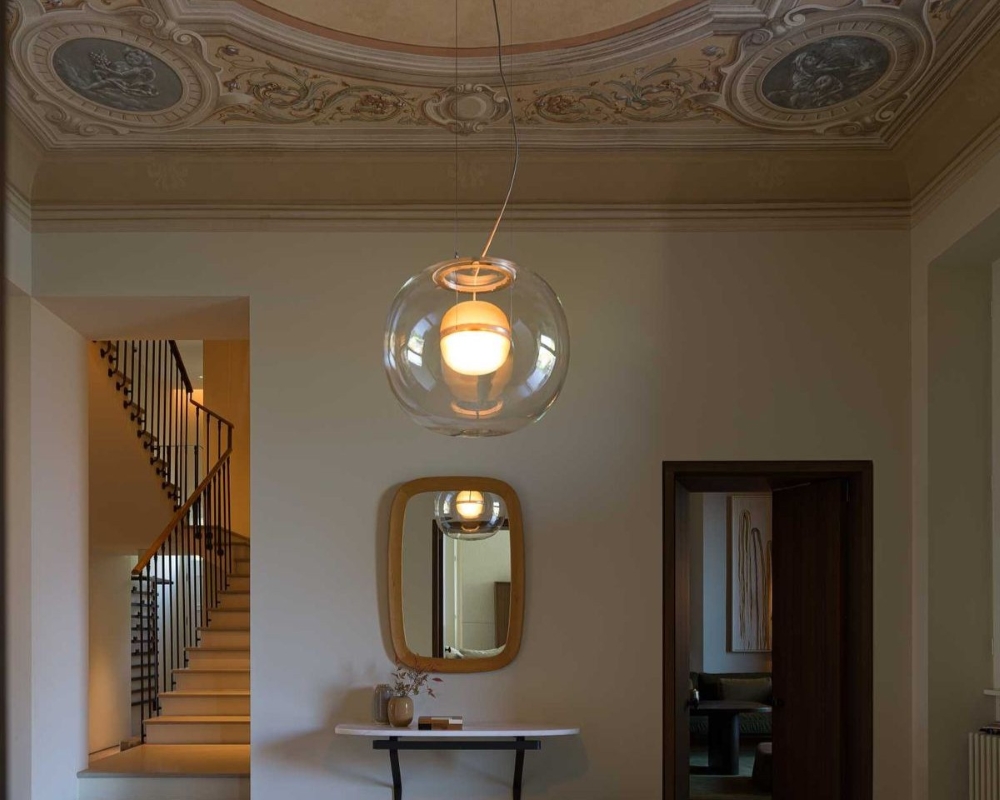 A corridor with a decorative painted ceiling, a Brokis glass pendant light, a console table with a vase, a round mirror on the wall, a staircase to the left, and a doorway leading to another room on the right.