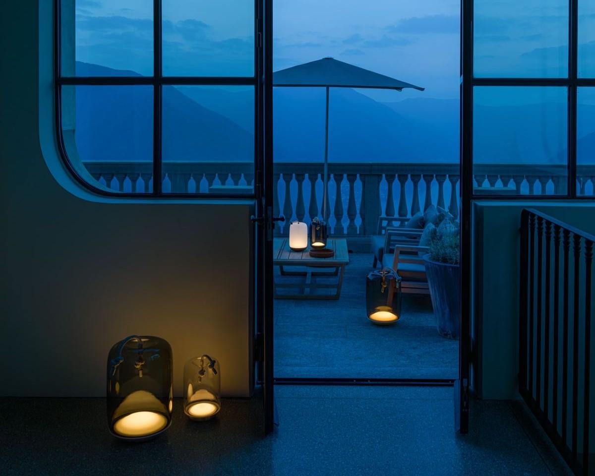A cosy terrace at dusk, viewed through open glass doors. Warm Brokis lanterns softly illuminate the floors inside and outside, with a table, chairs, and umbrella on the balcony against a backdrop of blue mountains.