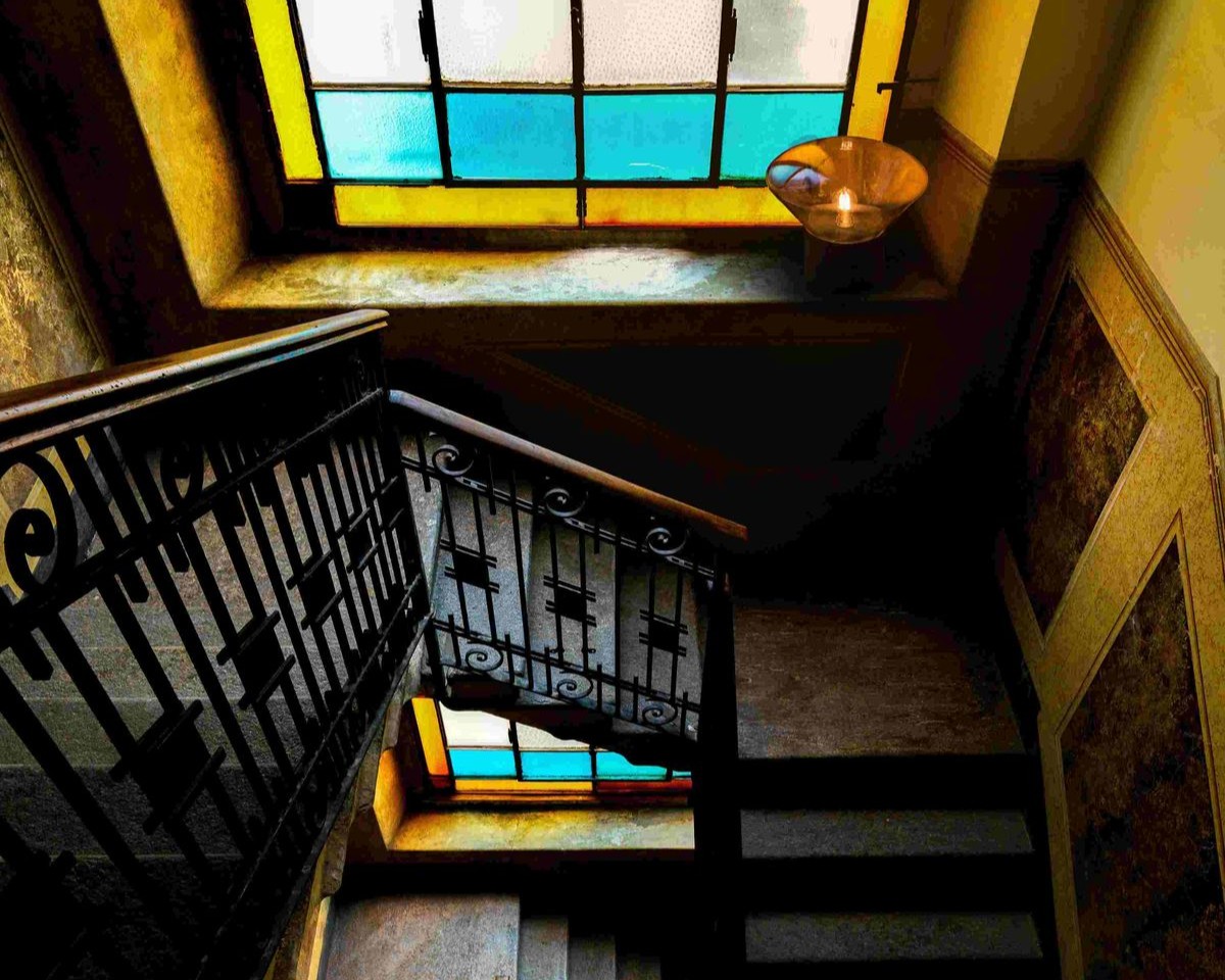 A view looking up a dimly lit stairwell with an ornate black balustrade, illuminated by stained glass windows with blue and yellow sections and a vintage Brokis ceiling light above.