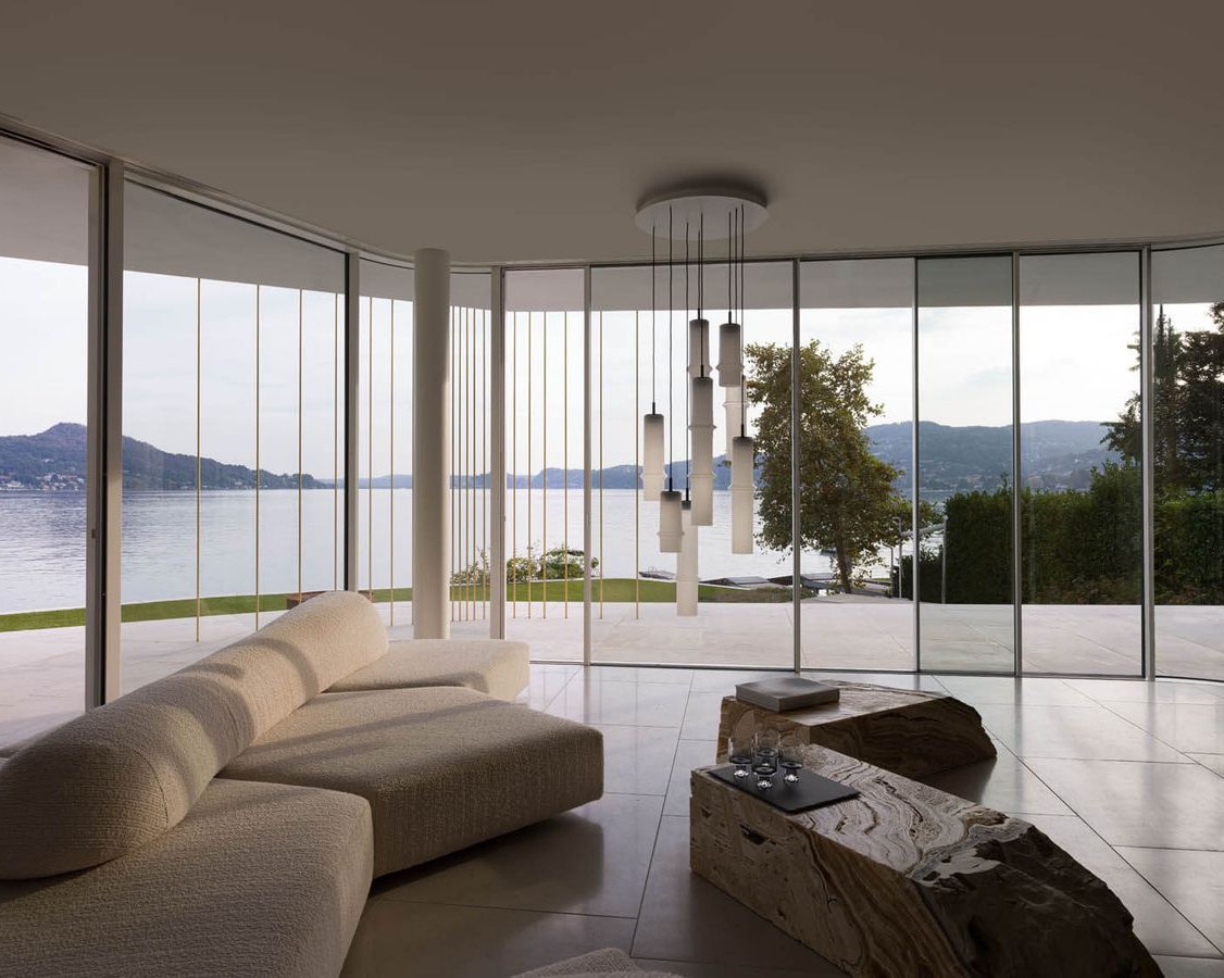 A modern living room with large floor-to-ceiling windows overlooking a lake and mountains. The room features a light curved sofa, unique wooden coffee tables, and Brokis pendant lights clustered elegantly from the ceiling.